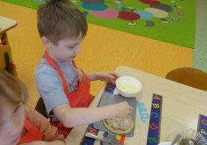 Dwójka dzieci dekoruje szynką, warzywami i serem placek w kształcie koła.